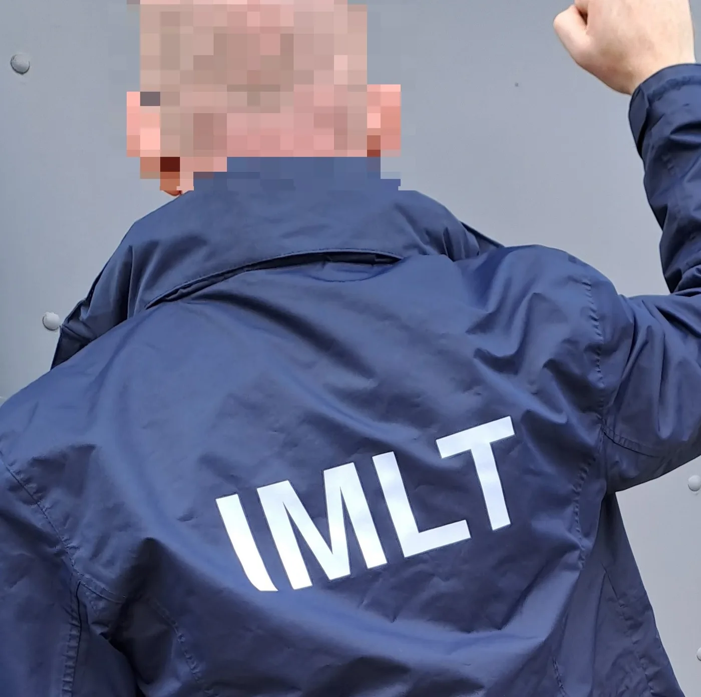The back of an officer's navy jacket printed with the letters IMLT, face pixelated